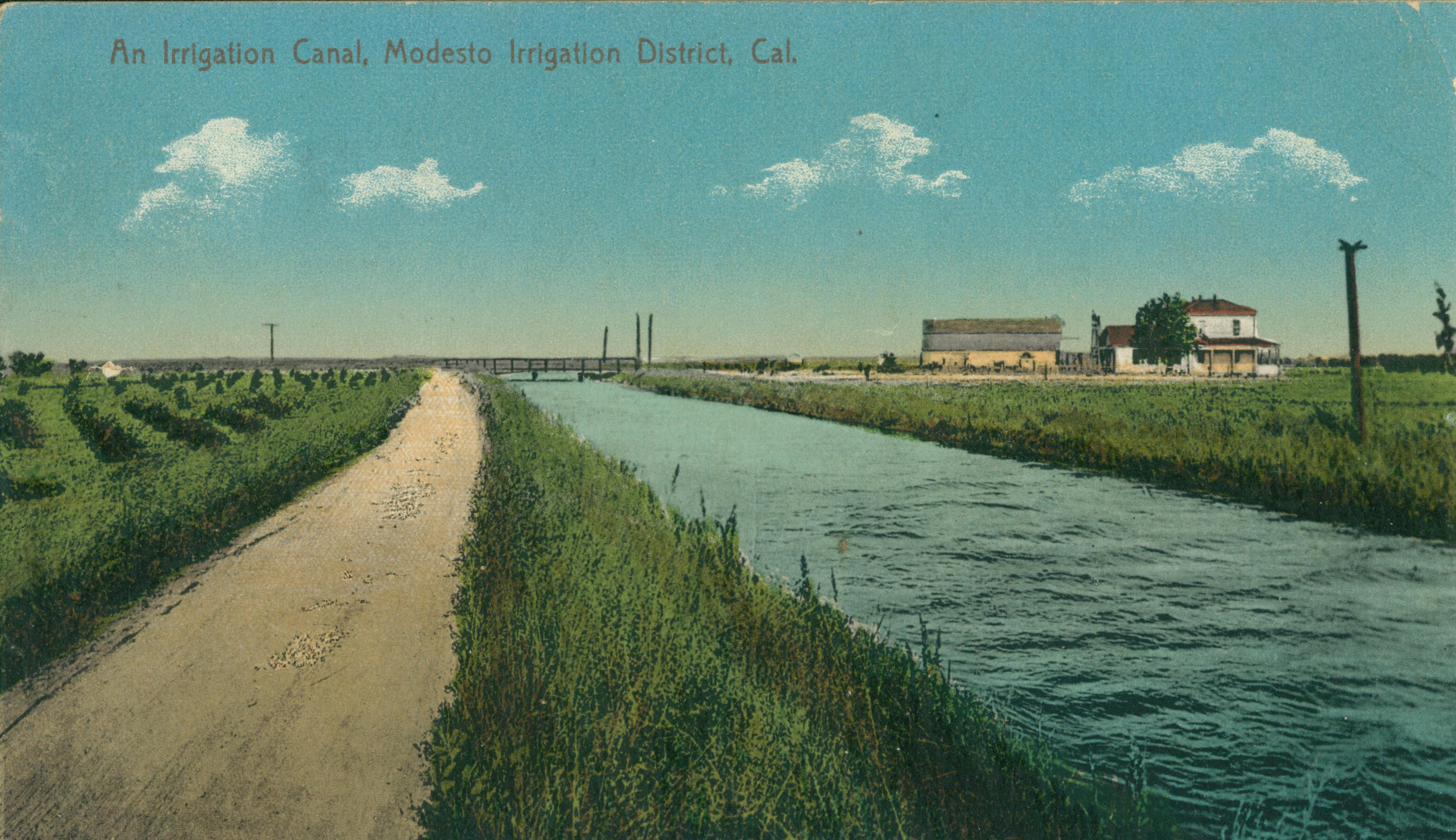 Shows an irrigation canal with a road to one side and farm buildings to the other